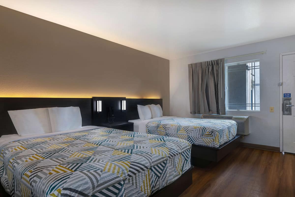 A hotel room with two beds, a large window with curtains, a wall-mounted air conditioner, and a door with a sign.