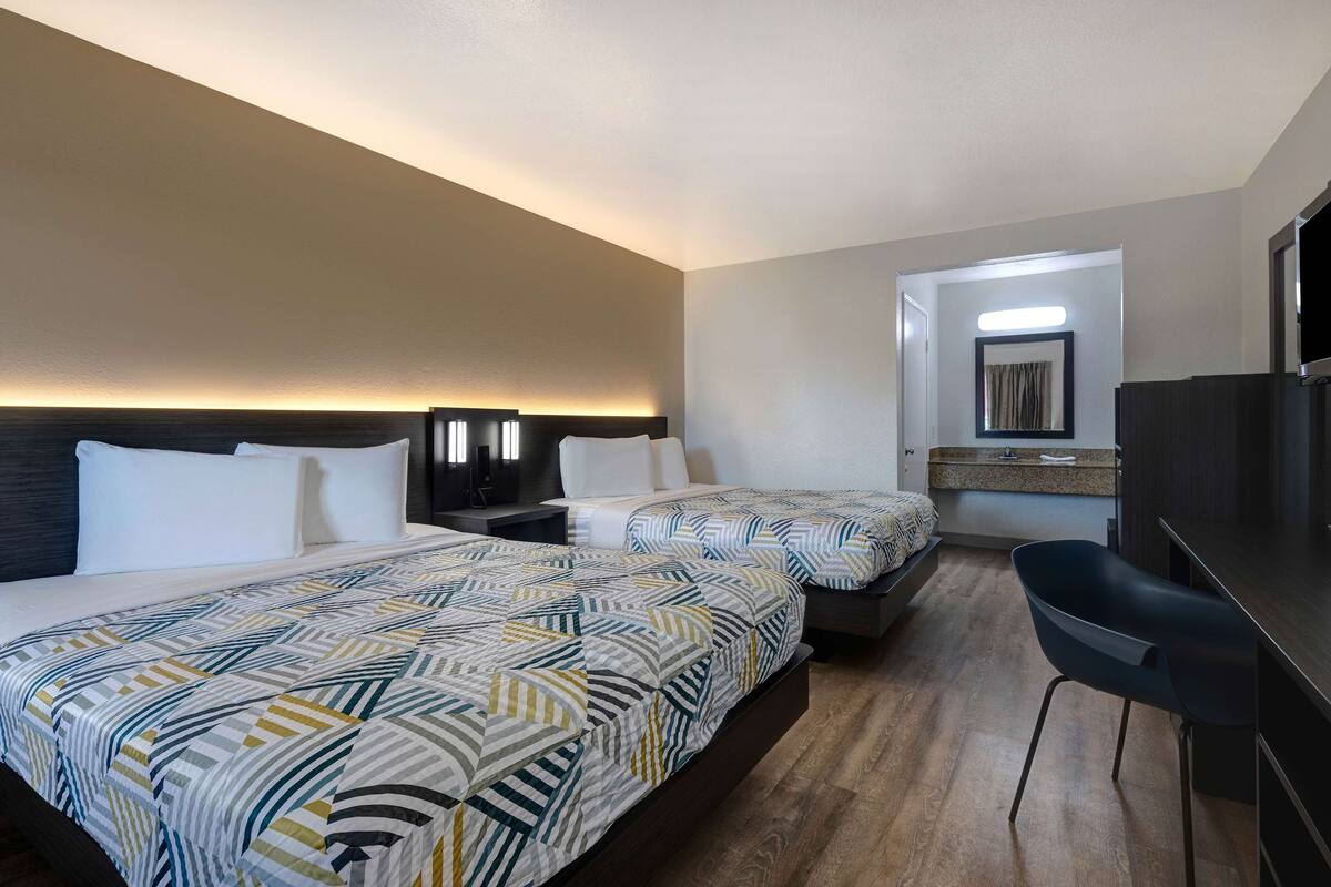 A hotel room with two beds, a desk with a chair, a flat-screen TV, and a bathroom visible through an open door.