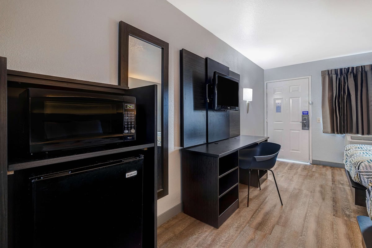 A compact hotel room with a kitchenette, a flat-screen TV, a bed, and a window with curtains.