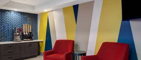 A modern hotel lobby with a bar area, red armchairs, and a colorful geometric wall design.