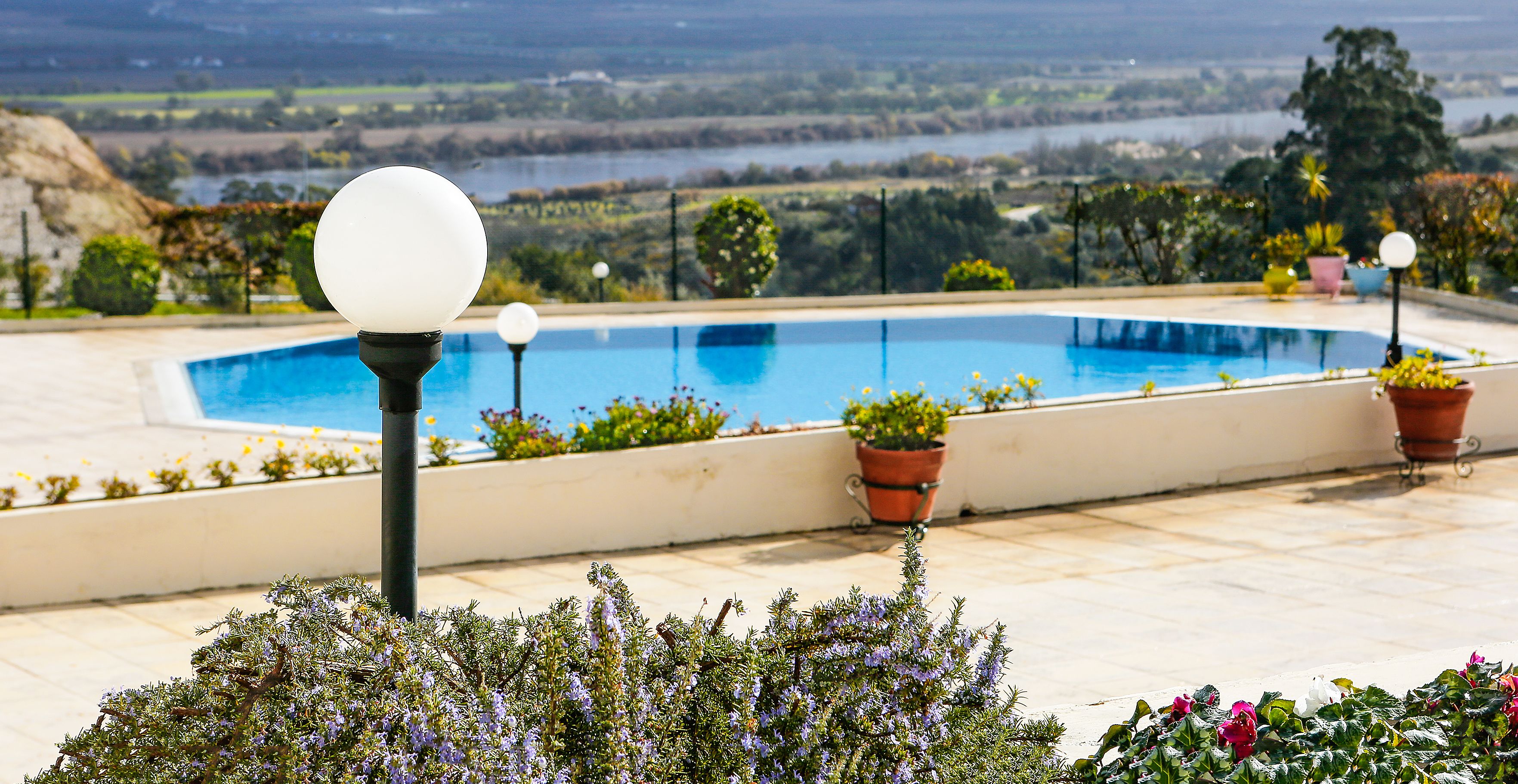 A swimming pool with a view of a lake and hills, surrounded by potted plants and a lamp post.