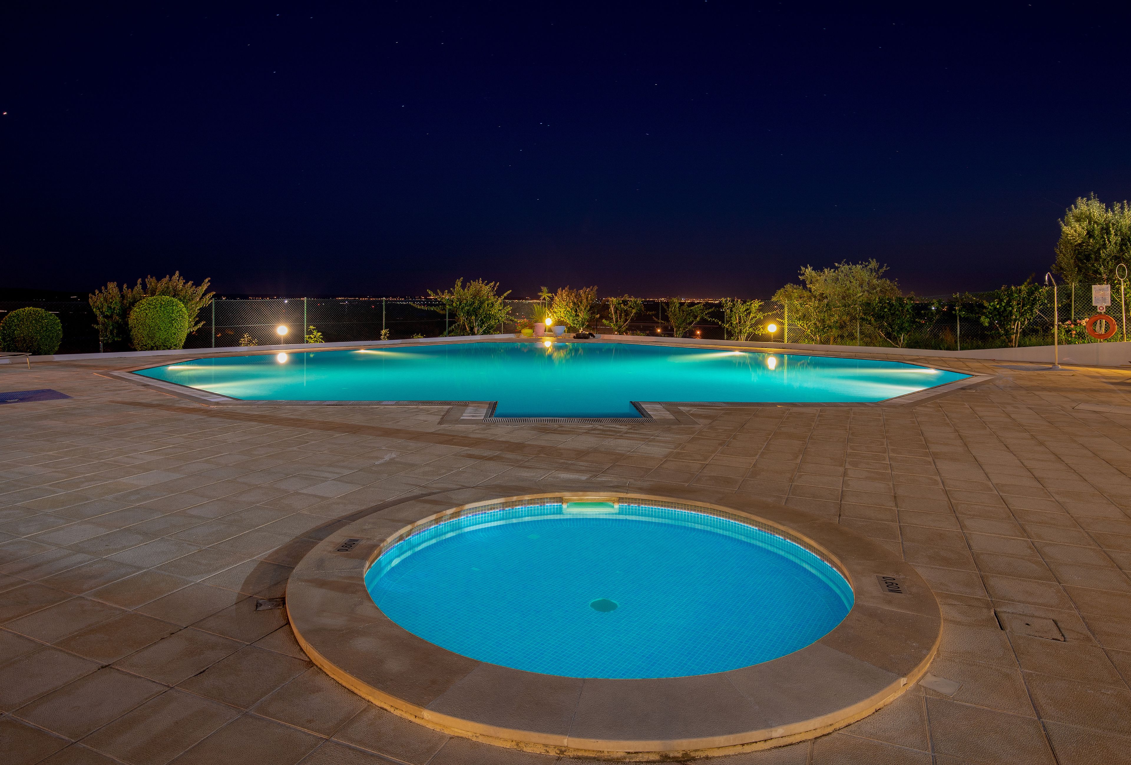 Une piscine avec un jacuzzi circulaire au centre, entourée d’une terrasse dallée et éclairée par des jeux de lumière la nuit.