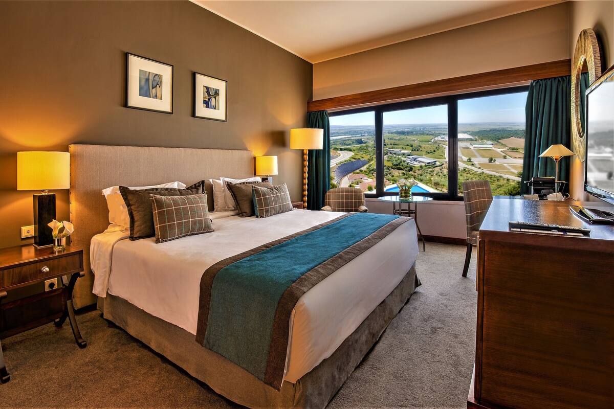 A hotel room with a large bed, two bedside lamps, a desk with a chair, a TV, and a view of a landscape through large windows.