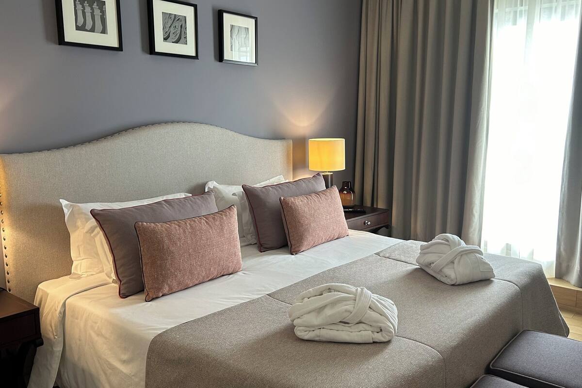 A hotel room with a large bed, multiple pillows, a bedside table with a lamp, and a window with curtains.