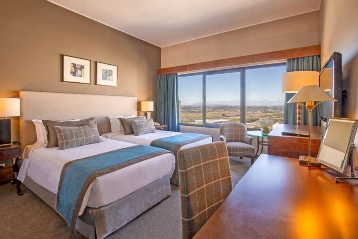 A hotel room with two beds, a desk, a chair, and a large window offering a view of the landscape.