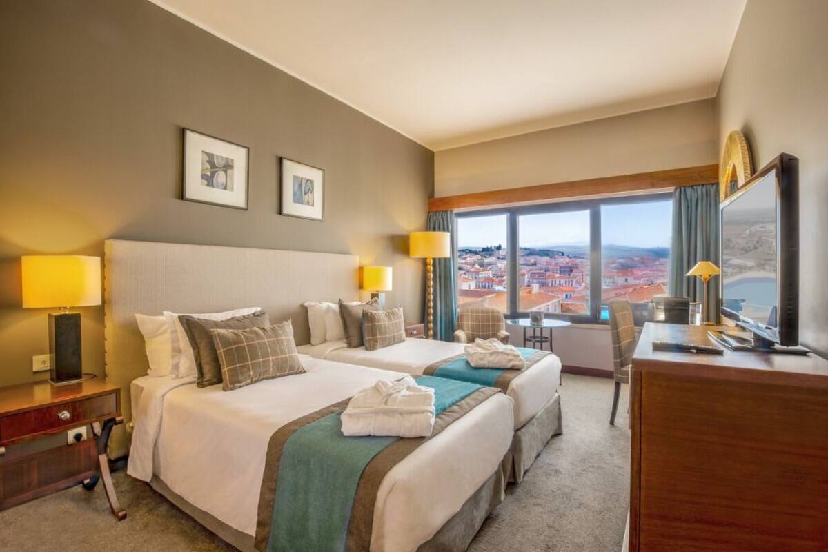 A hotel room with two beds, a desk, a chair, and a large window offering a city view.