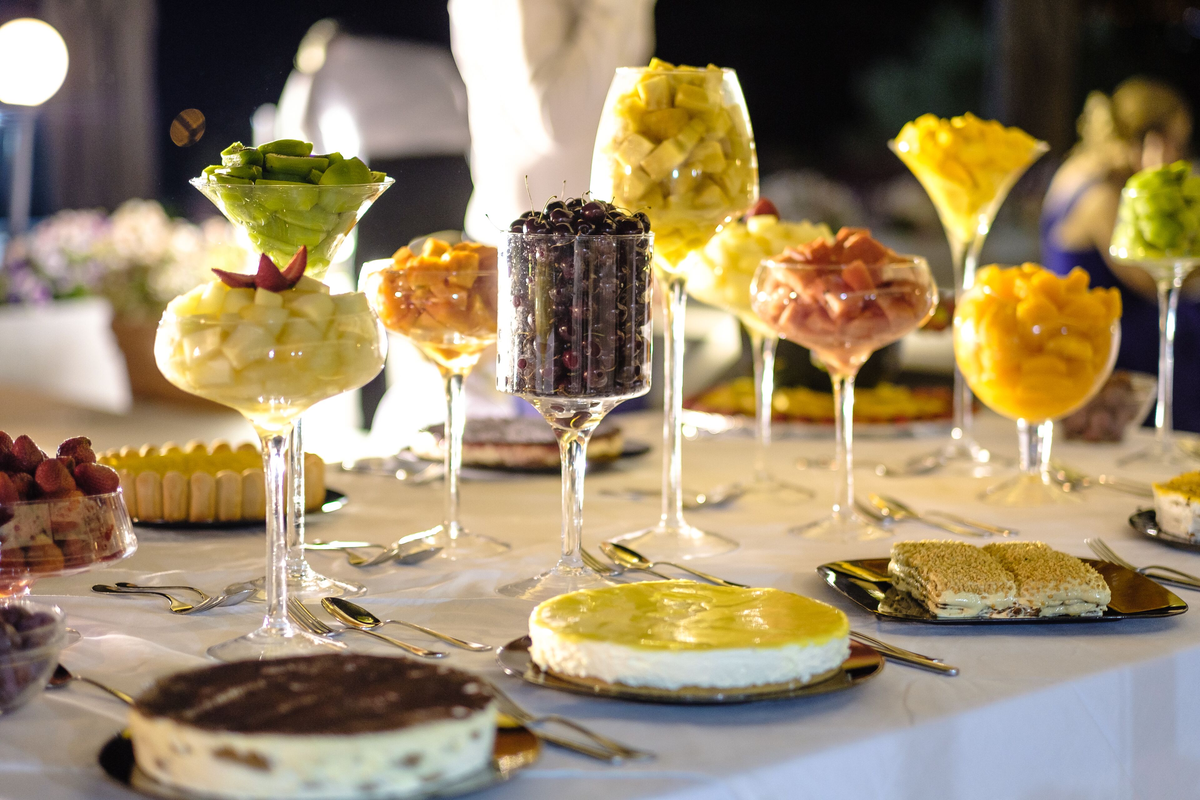 A table with various desserts and drinks, including a cheesecake, a fruit salad, and a glass of wine.