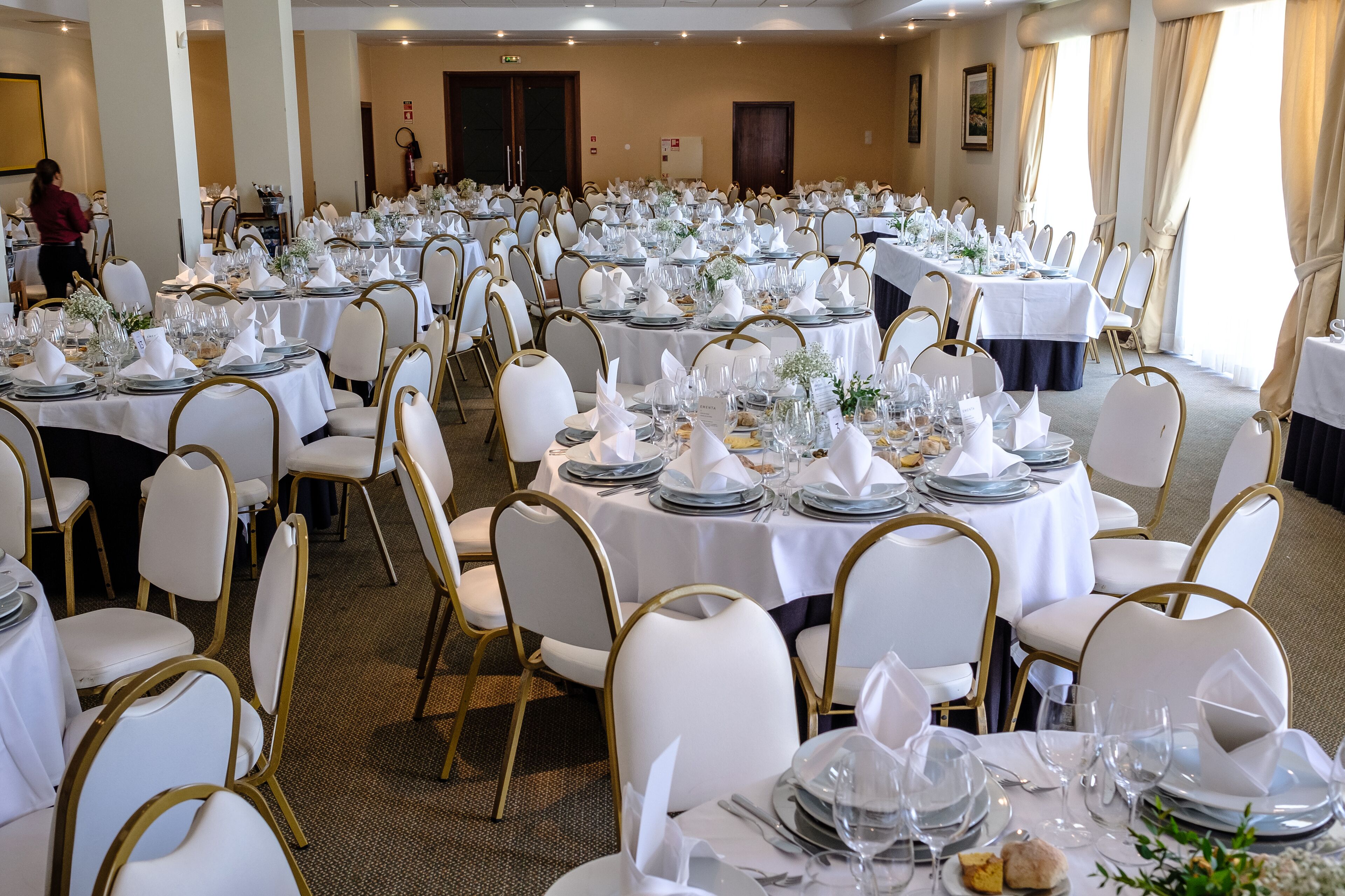 A banquet hall with round tables set for a formal event, featuring white tablecloths, gold chairs, and neatly arranged table settings.