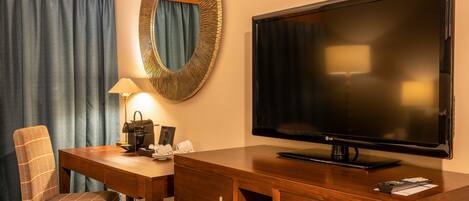 A hotel room with a large flat-screen TV, a wooden desk, a chair, a mirror, and a lamp.