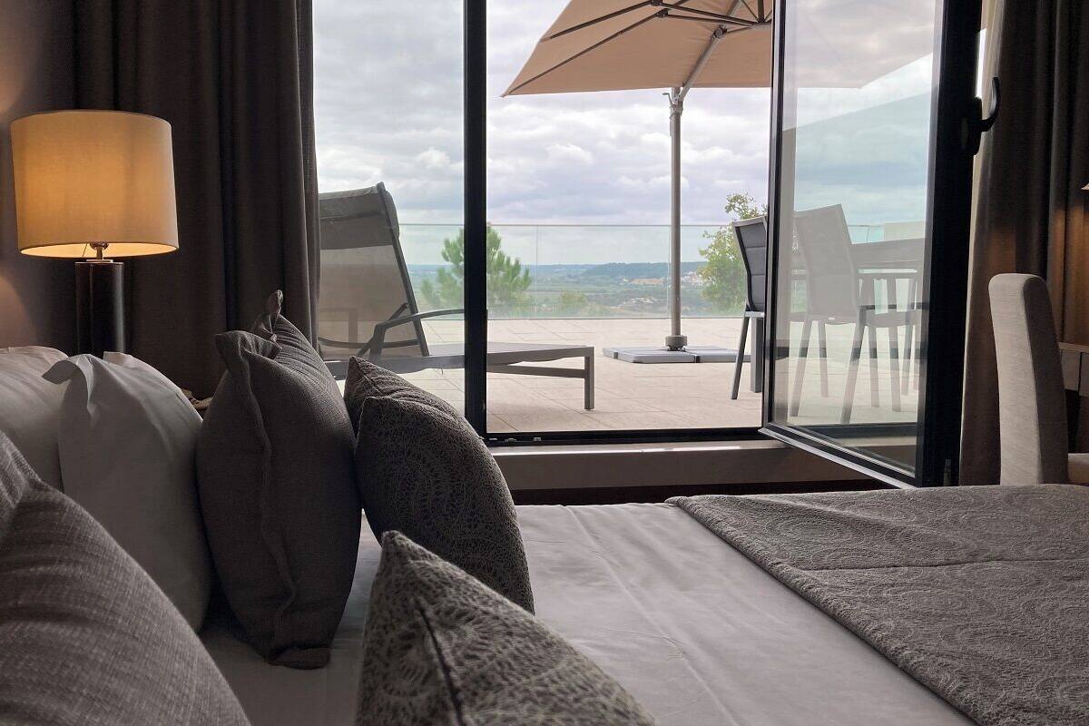 A hotel room with a bed, pillows, a lamp, and a view of the outdoors through a large window.