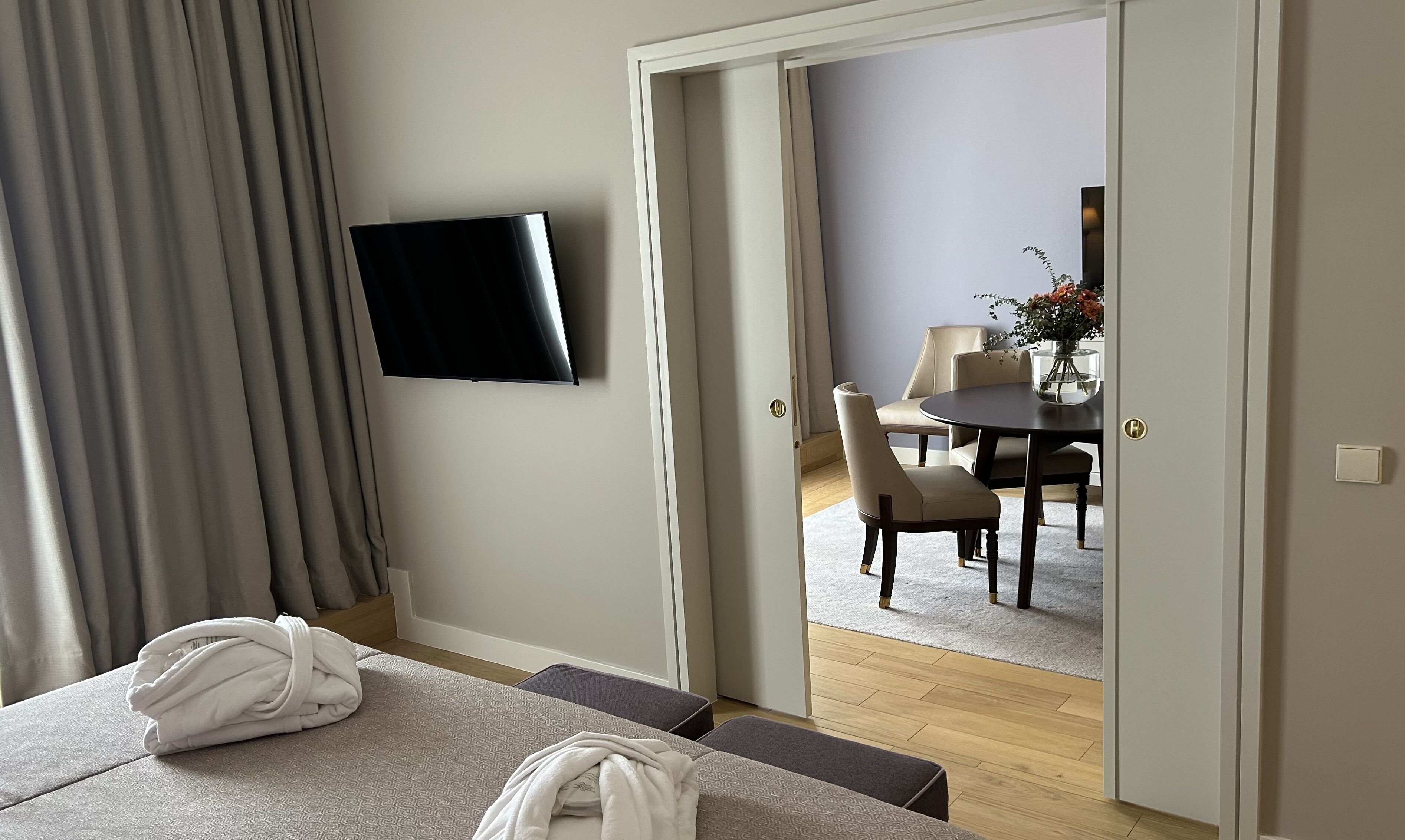 Hotellihuone, jossa on sänky, seinälle asennettu televisio, ruokailutila pöytineen ja tuoleineen sekä ovi, joka johtaa toiseen huoneeseen.