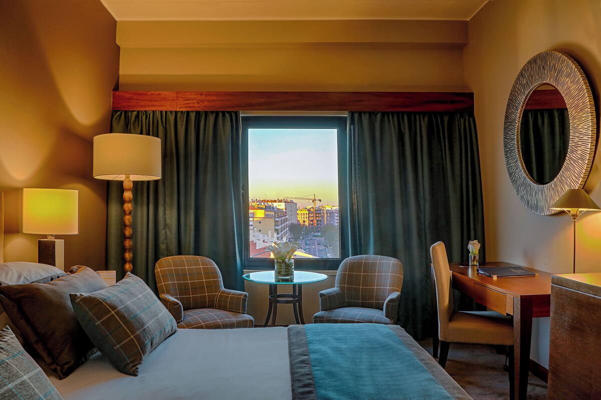 A hotel room with a bed, two chairs, a desk, a lamp, a mirror, and a view of a cityscape through a window.