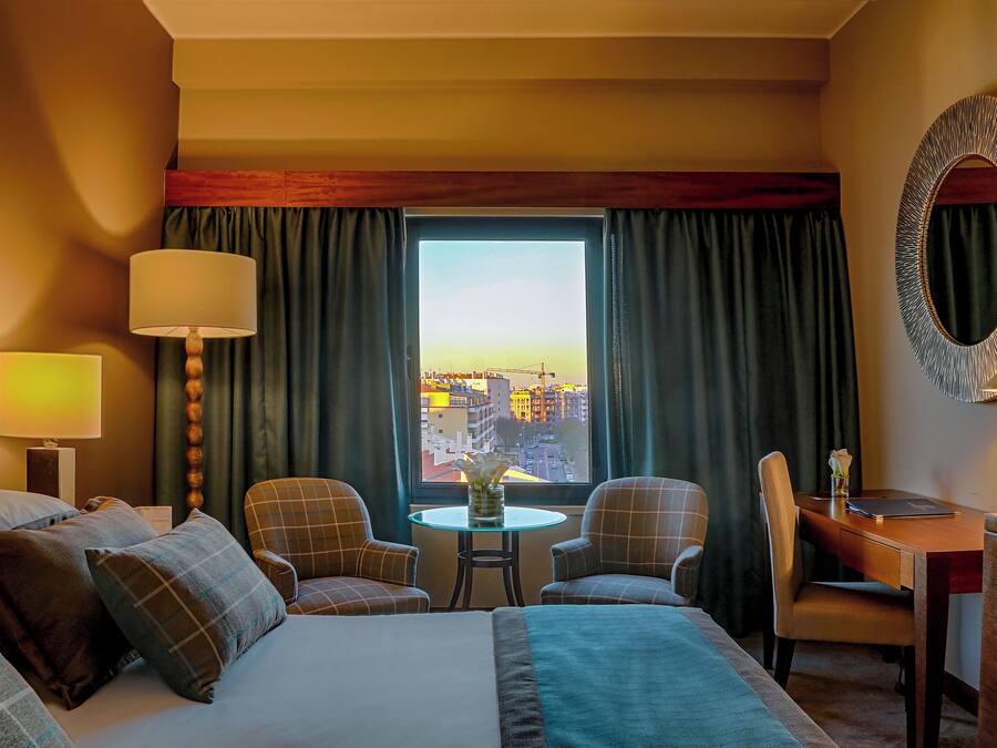 A hotel room with a bed, two chairs, a desk, a lamp, a mirror, and a view of a cityscape through a window.