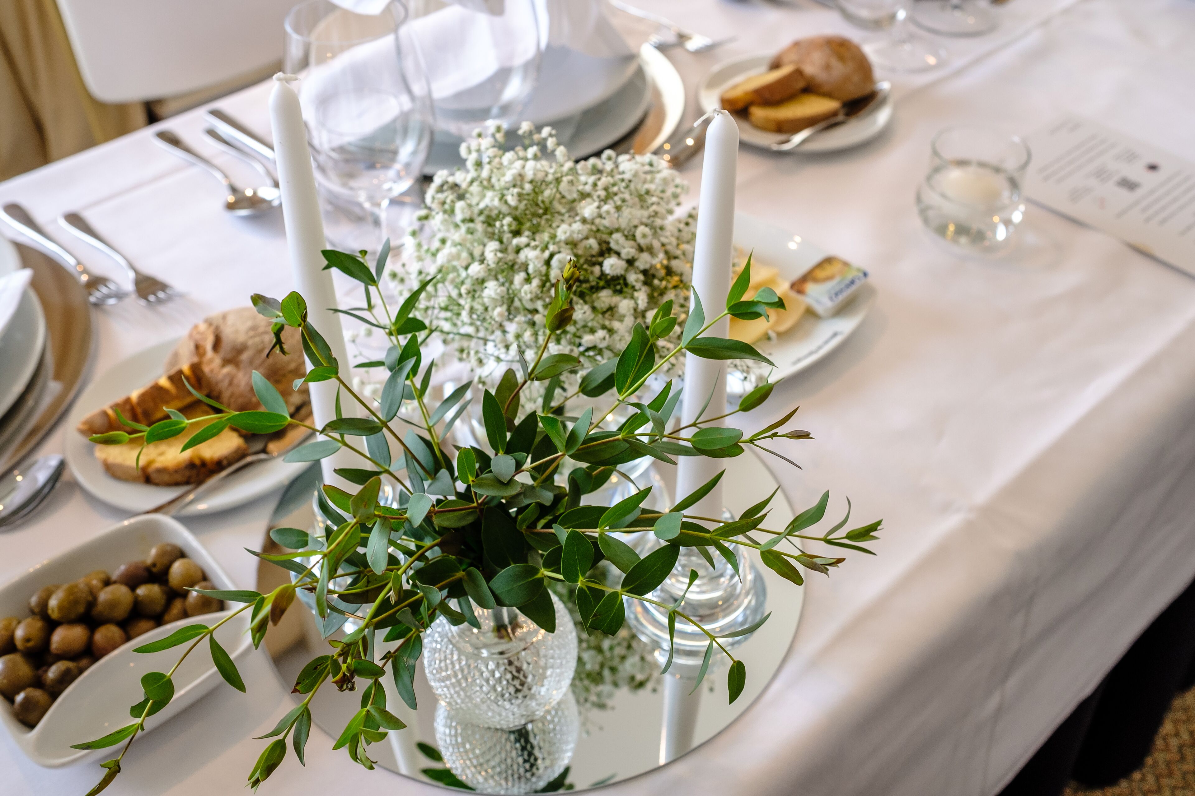 Una mesa dispuesta con platos blancos, cubiertos y un centro de mesa con follaje verde y flores blancas.
