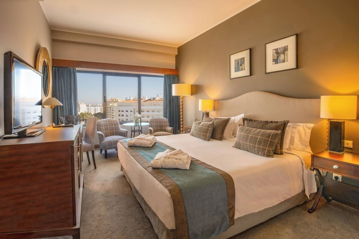 A hotel room with a large bed, two bedside lamps, a desk with a chair, a TV, and a view of buildings through the window.
