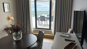 Una habitación de hotel con una mesa redonda, un florero con flores, una silla, un sofá, un televisor y vistas al exterior a través de un ventanal grande.
