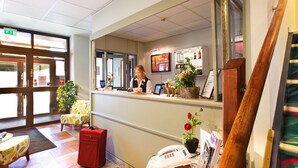 Un hall d’hôtel avec un comptoir d’accueil, une réceptionniste, une valise rouge, un téléphone et une plante.