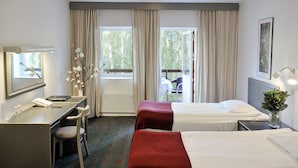 Une chambre d’hôtel avec deux lits, un bureau, une chaise, une lampe de bureau, un vase avec des fleurs et une fenêtre avec des rideaux.