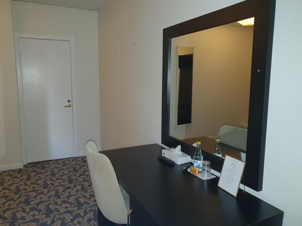 A hotel room with a large mirror, a desk with a chair, a bottle of water, a tissue box, and a sign.