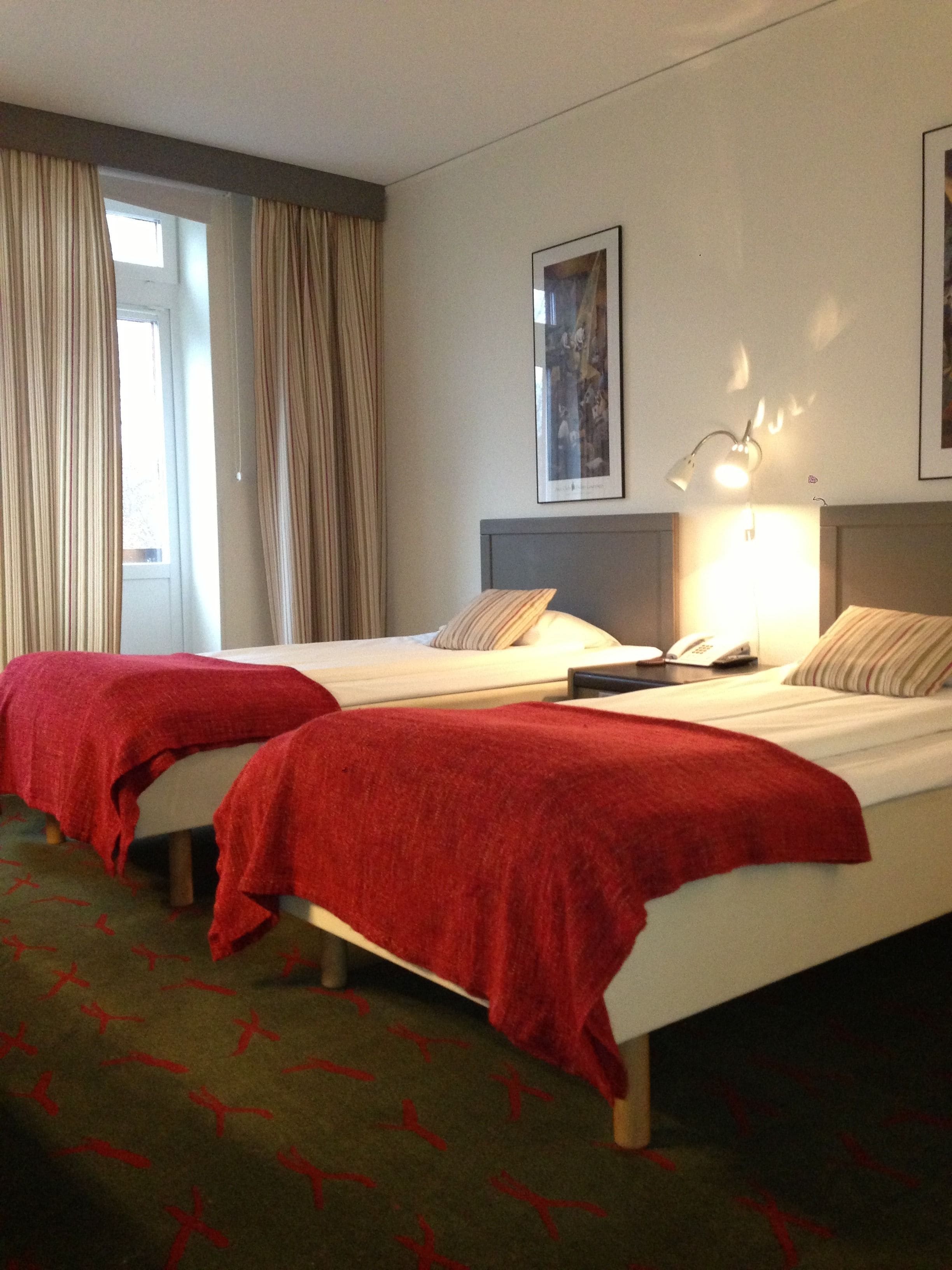 A hotel room with two beds, a large window with curtains, a framed picture on the wall, and a bedside lamp.