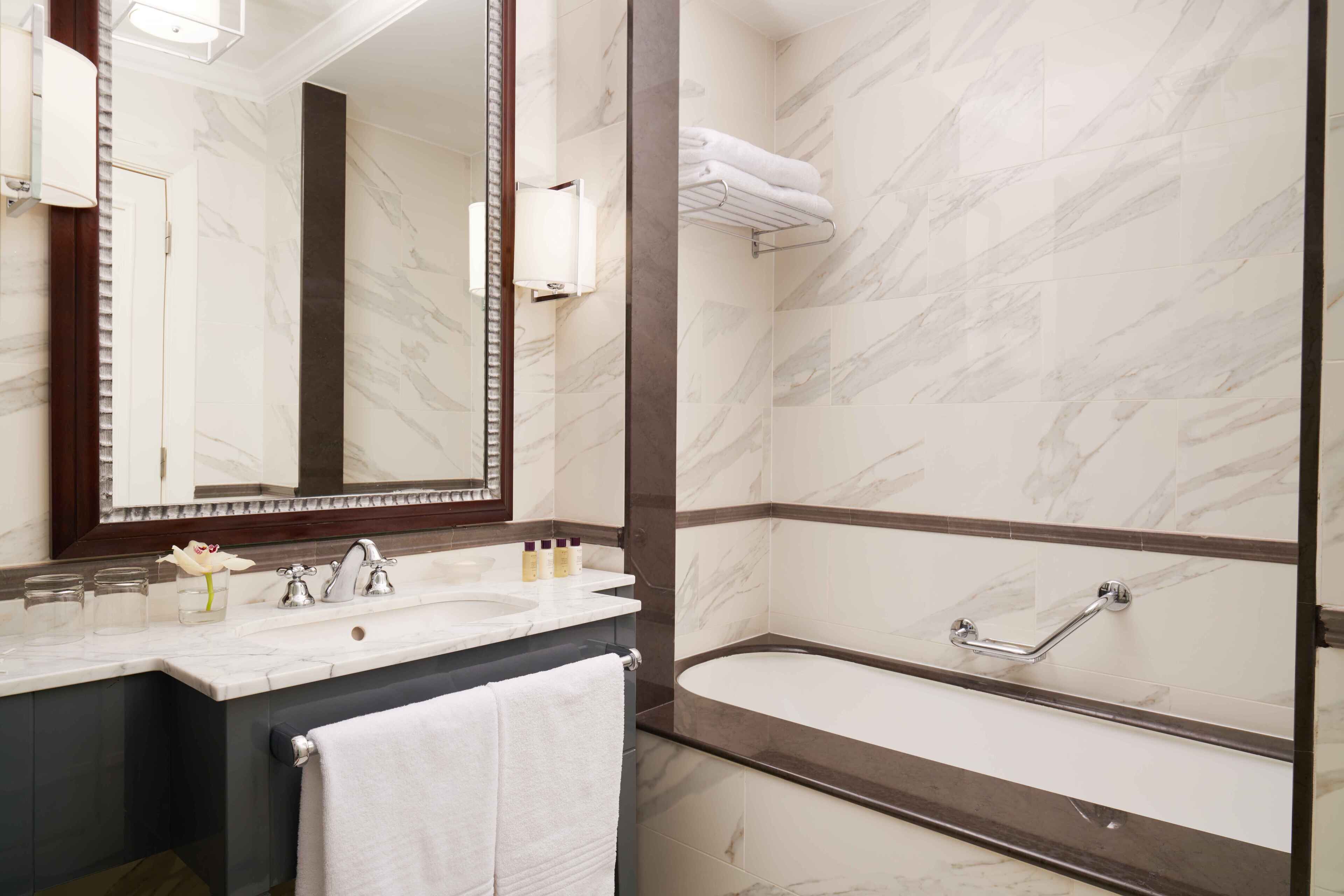 A modern bathroom with a marble vanity, a large mirror, a bathtub, and a towel rack.
