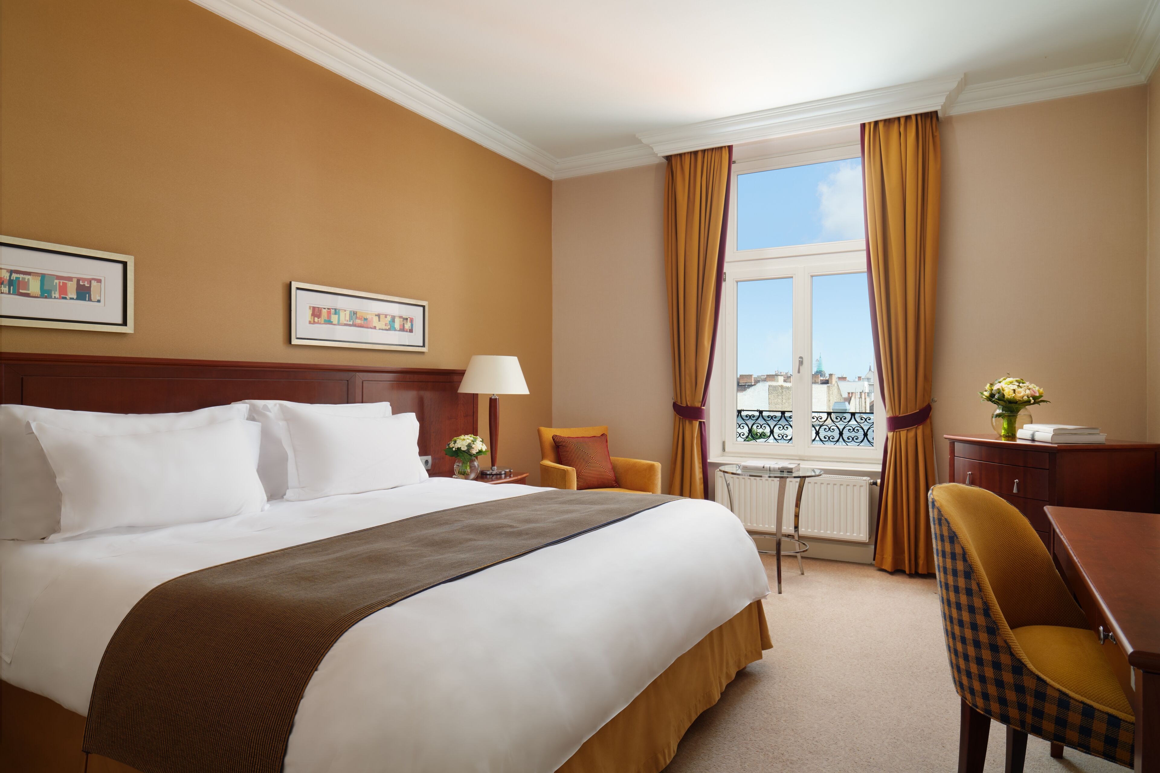 A hotel room with a large bed, a desk, a chair, a window with curtains, and framed pictures on the wall.