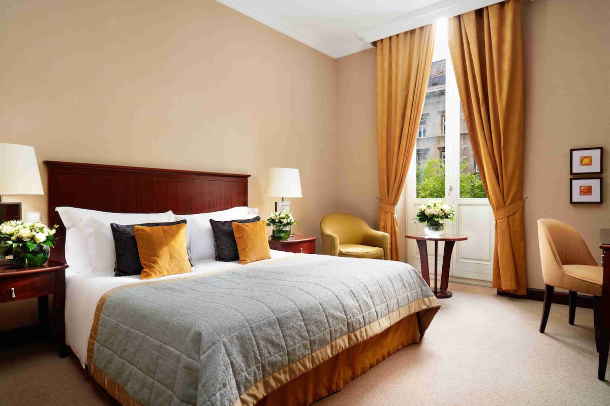 Een hotelkamer met een groot bed, twee nachtkastjes met lampen, een stoel, een klein tafeltje met bloemen en een raam met gordijnen.