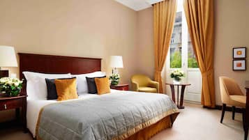 Habitació d'hotel amb un llit gran, dues taules de nit amb làmpades, una cadira, una taula petita amb flors i una finestra amb cortines.