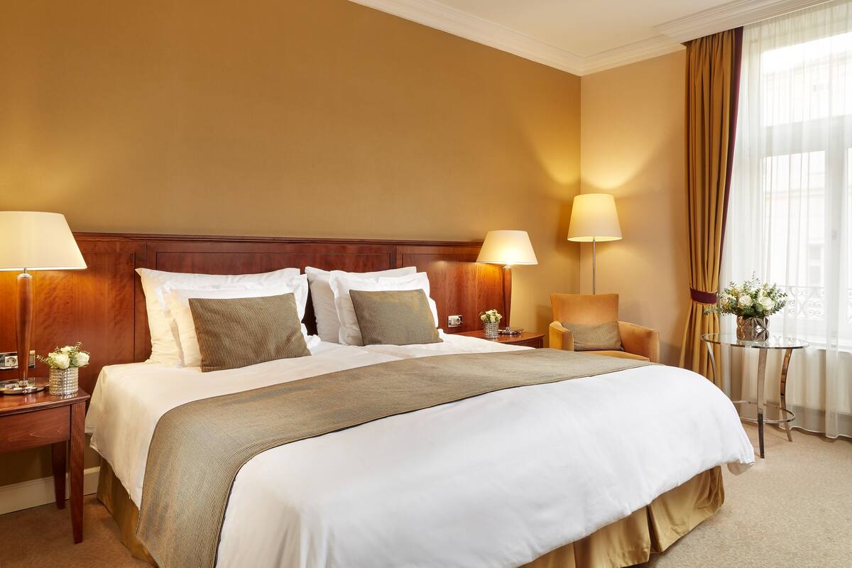Een hotelkamer met een groot bed, twee nachtkastjes met lampen, een tafeltje met een bloemenvaas, en een raam met gordijnen.