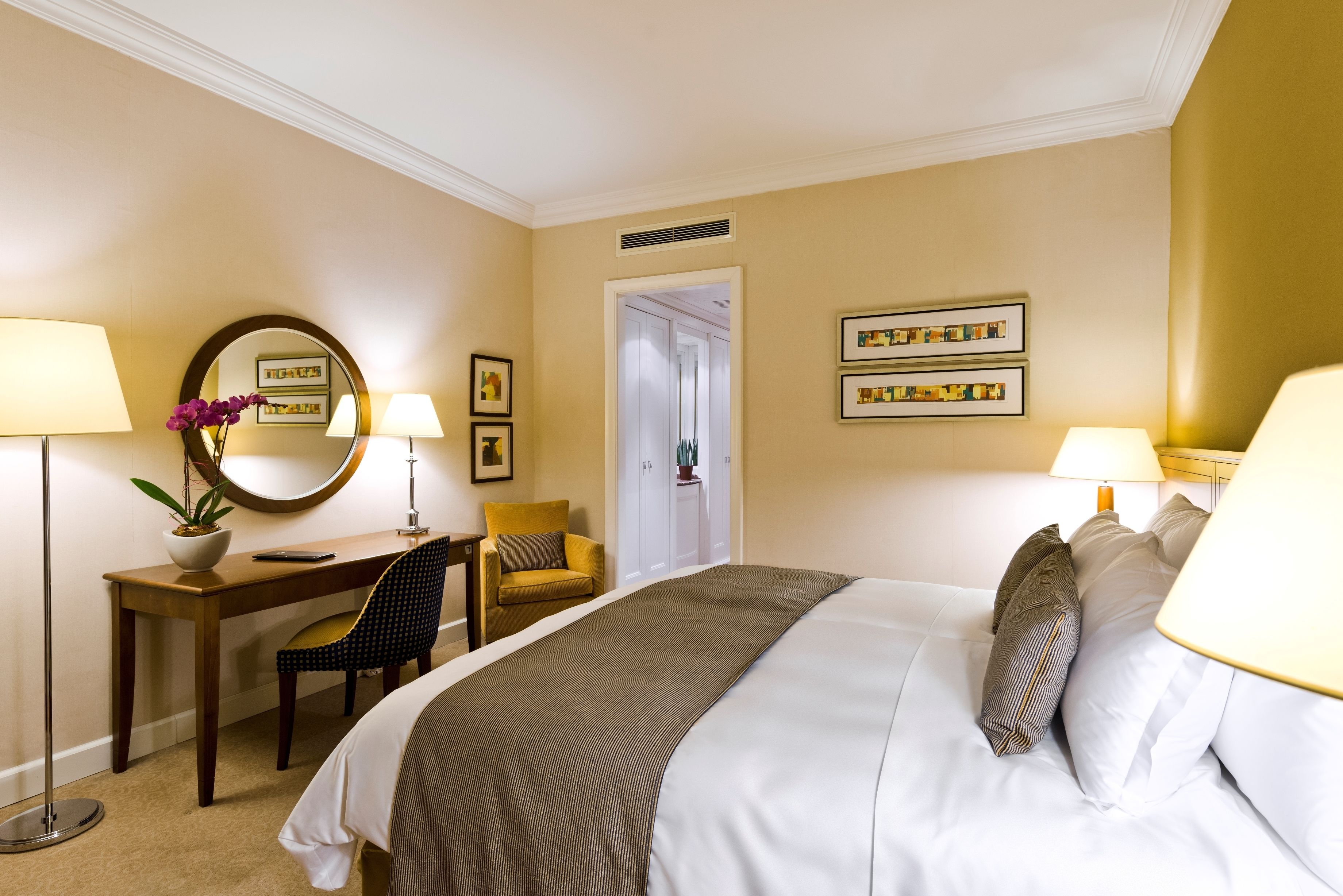A hotel room with a bed, two bedside lamps, a desk with a chair, a potted plant, and framed pictures on the wall.