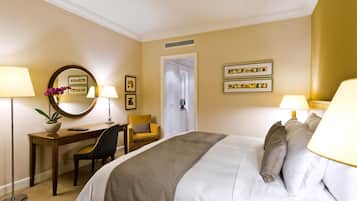 A hotel room with a bed, two bedside lamps, a desk with a chair, a potted plant, and framed pictures on the wall.