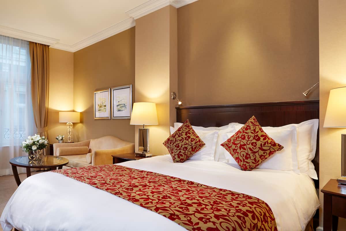 Hotelkamer met een groot bed, twee nachtkastjes, een stoel, een ronde tafel met een bloemenvaas en twee wandlampen.
