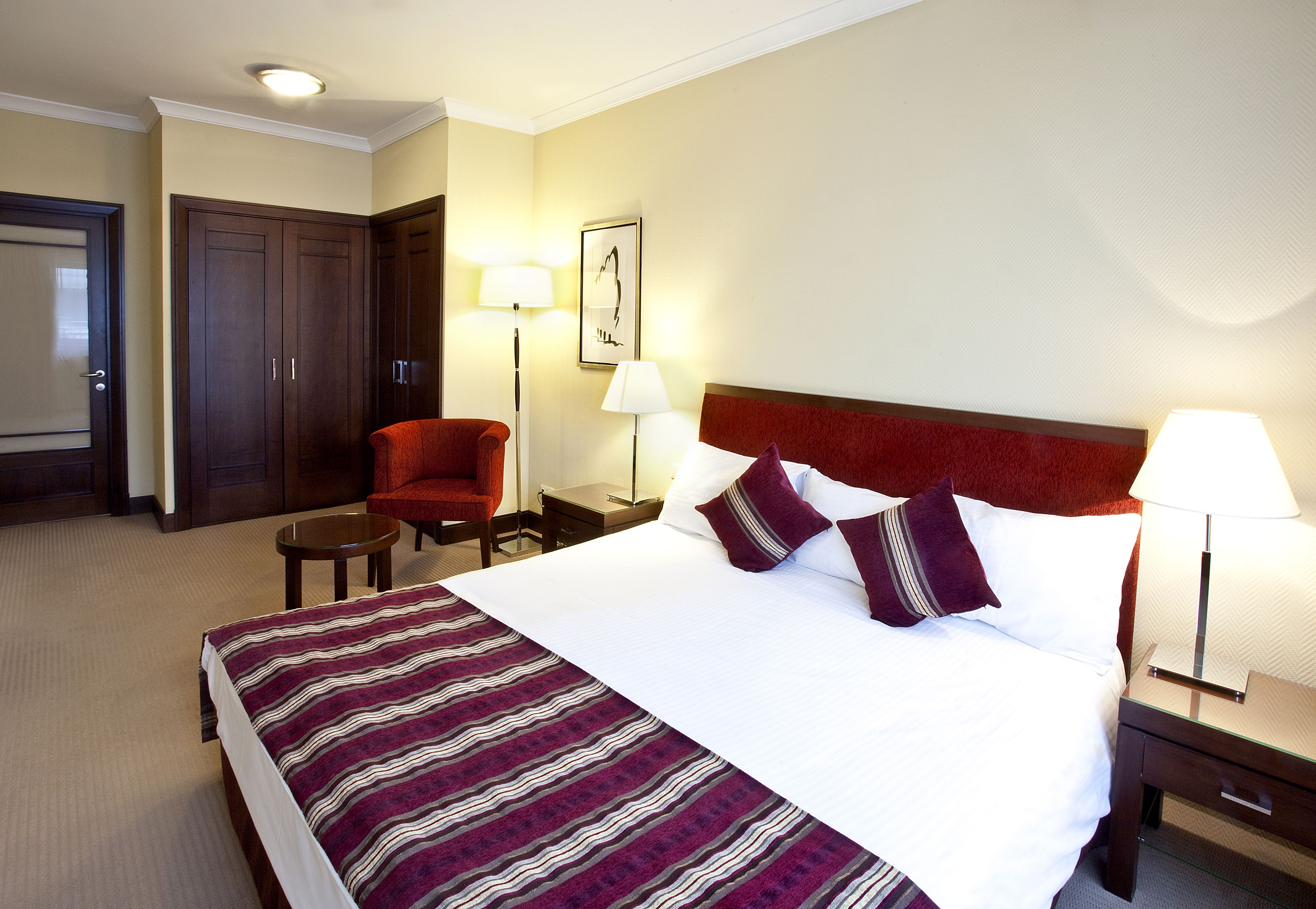 A hotel room with a large bed, two bedside tables with lamps, a red chair, and a wardrobe.