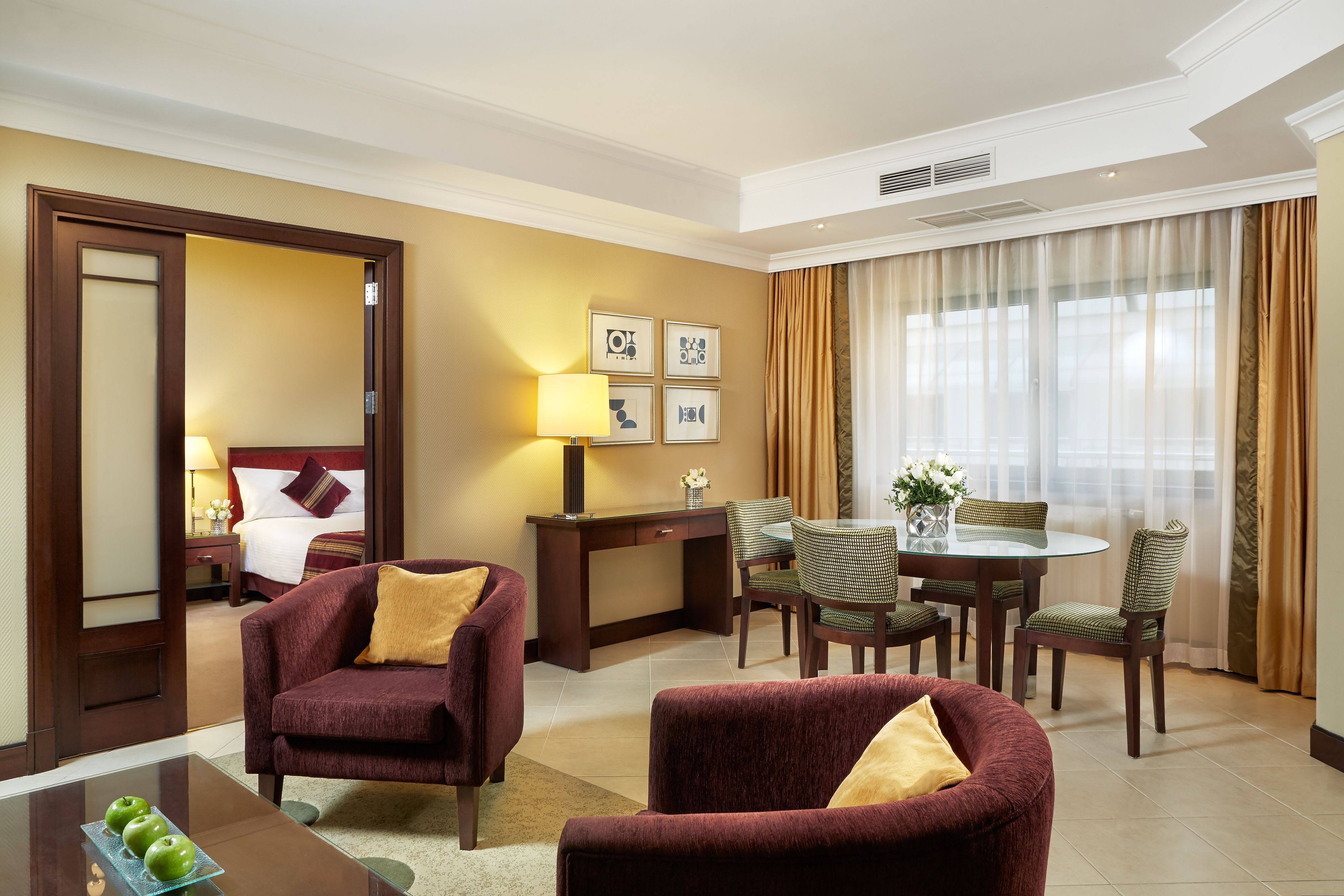 A hotel room with a bed, two armchairs, a desk, a dining table with chairs, and a large window with curtains.