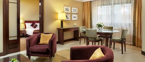 A hotel room with a bed, two armchairs, a desk, a dining table with chairs, and a large window with curtains.
