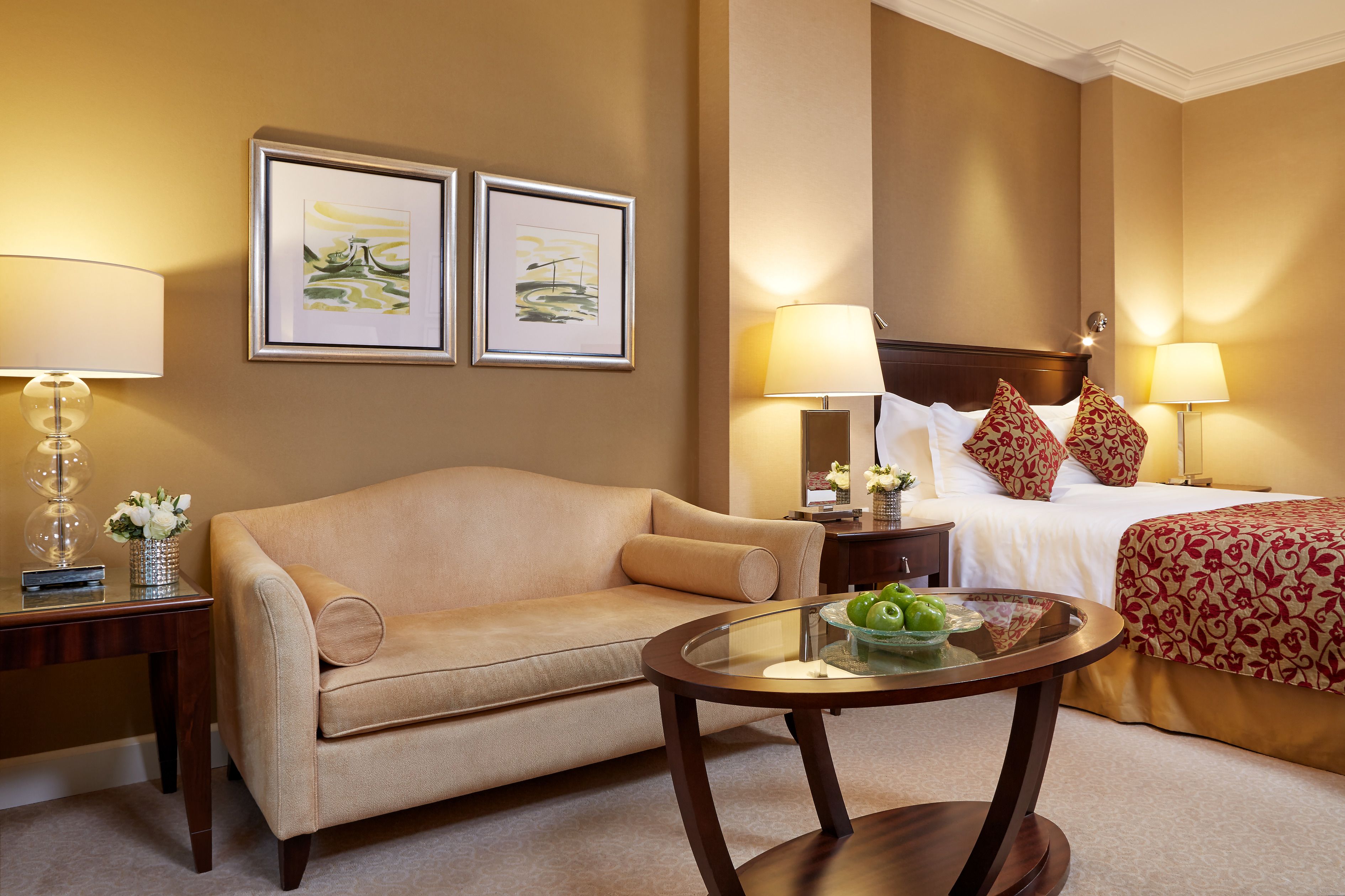A hotel room with a beige sofa, a round glass table with a green bowl, two framed artworks on the wall, a bed with white linens and red patterned pillows, and two bedside lamps.