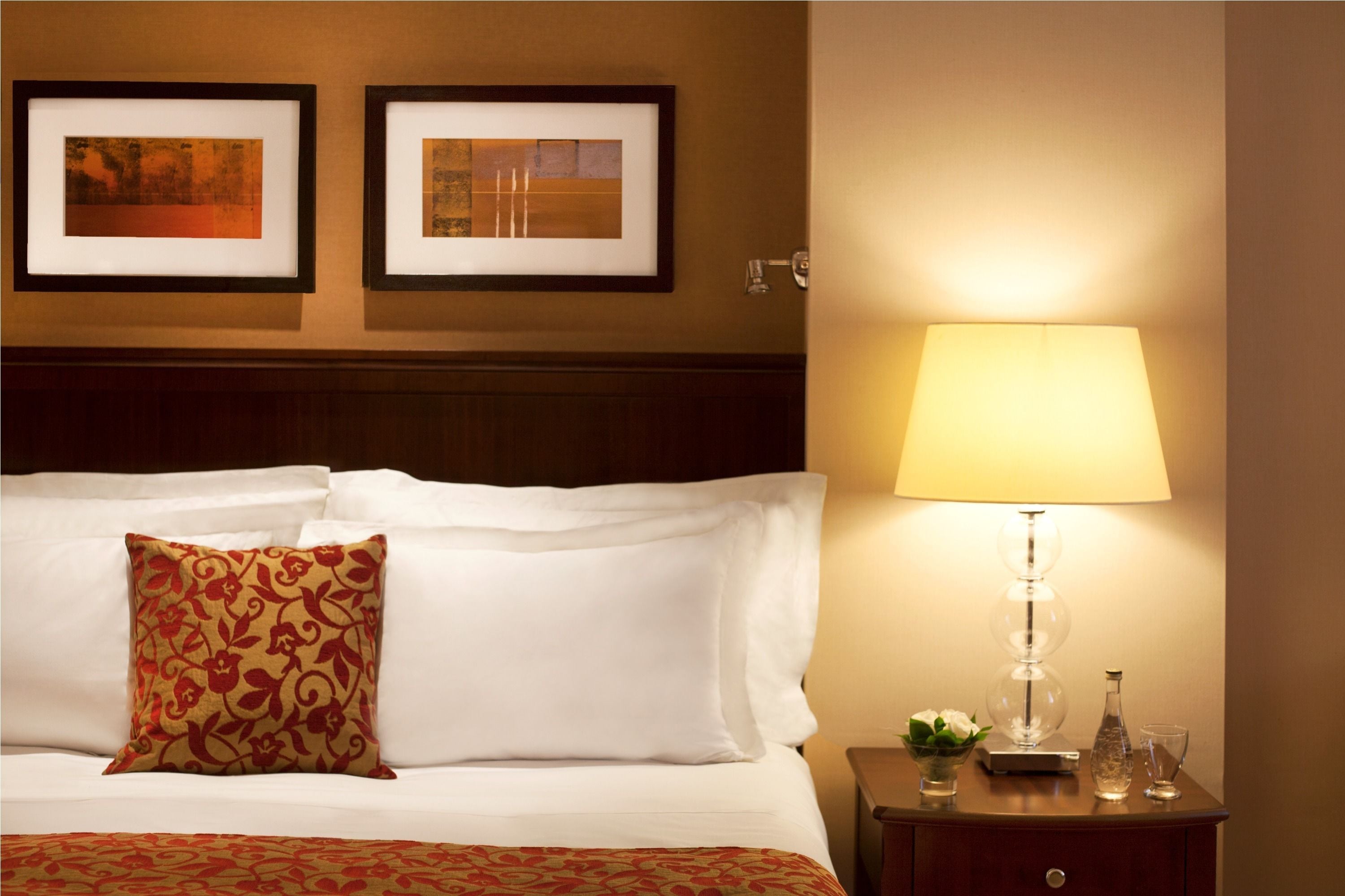 A neatly made bed with white linens, a patterned throw, and a single decorative pillow. A bedside table with a lamp, a glass, and a small vase is visible. Two framed artworks are hung on the wall above the bed.