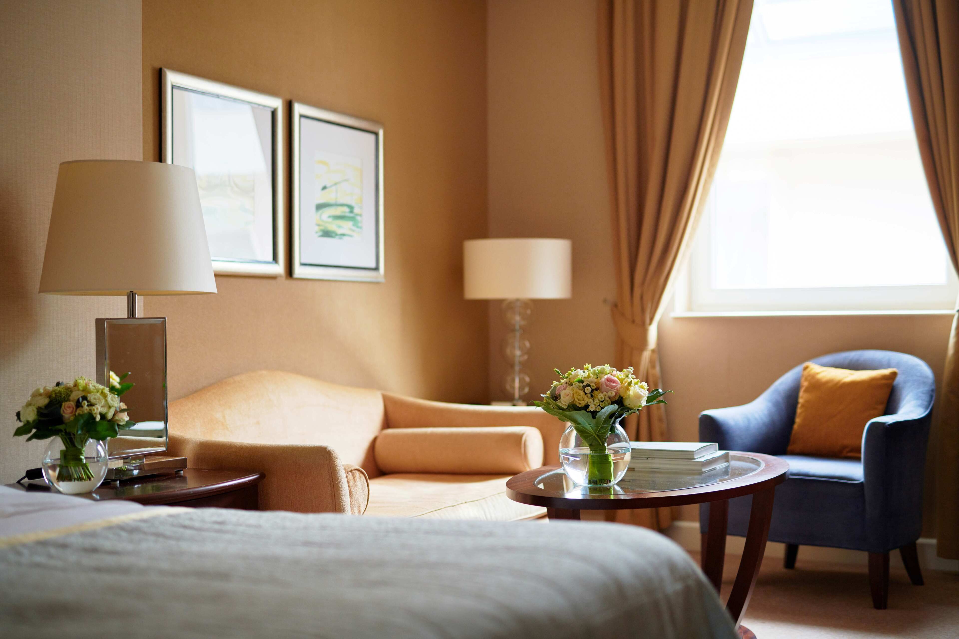 Habitación de hotel con cama, sofá, sillón azul, una mesa redonda con un jarrón de flores y dos cuadros en la pared.
