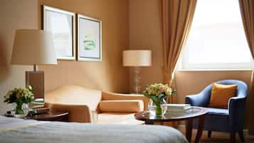 Habitación de hotel con cama, sofá, sillón azul, una mesa redonda con un jarrón de flores y dos cuadros en la pared.
