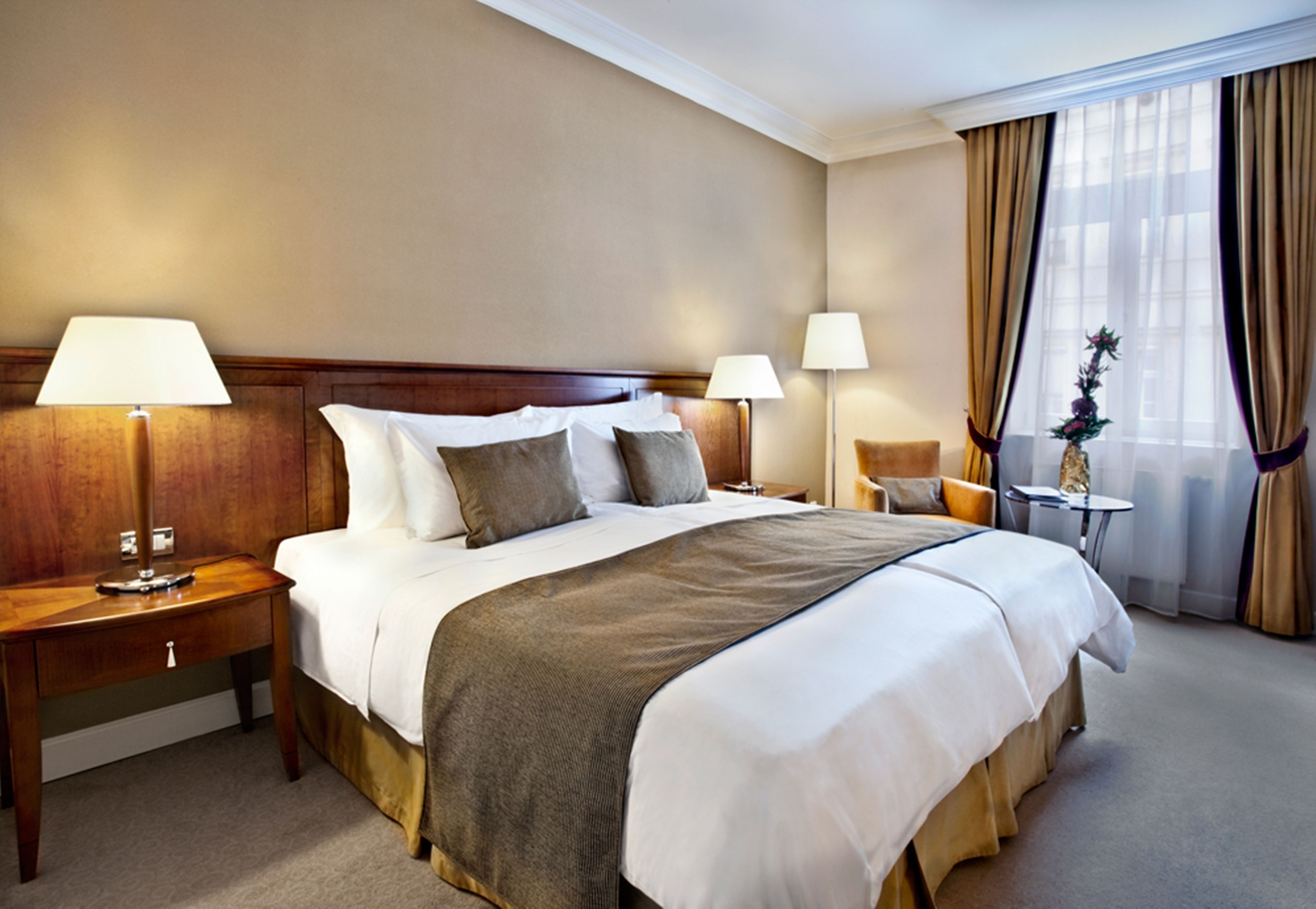 Habitación de hotel con una cama grande, dos mesitas de noche con lámparas, una mesita redonda pequeña y una ventana con cortinas.