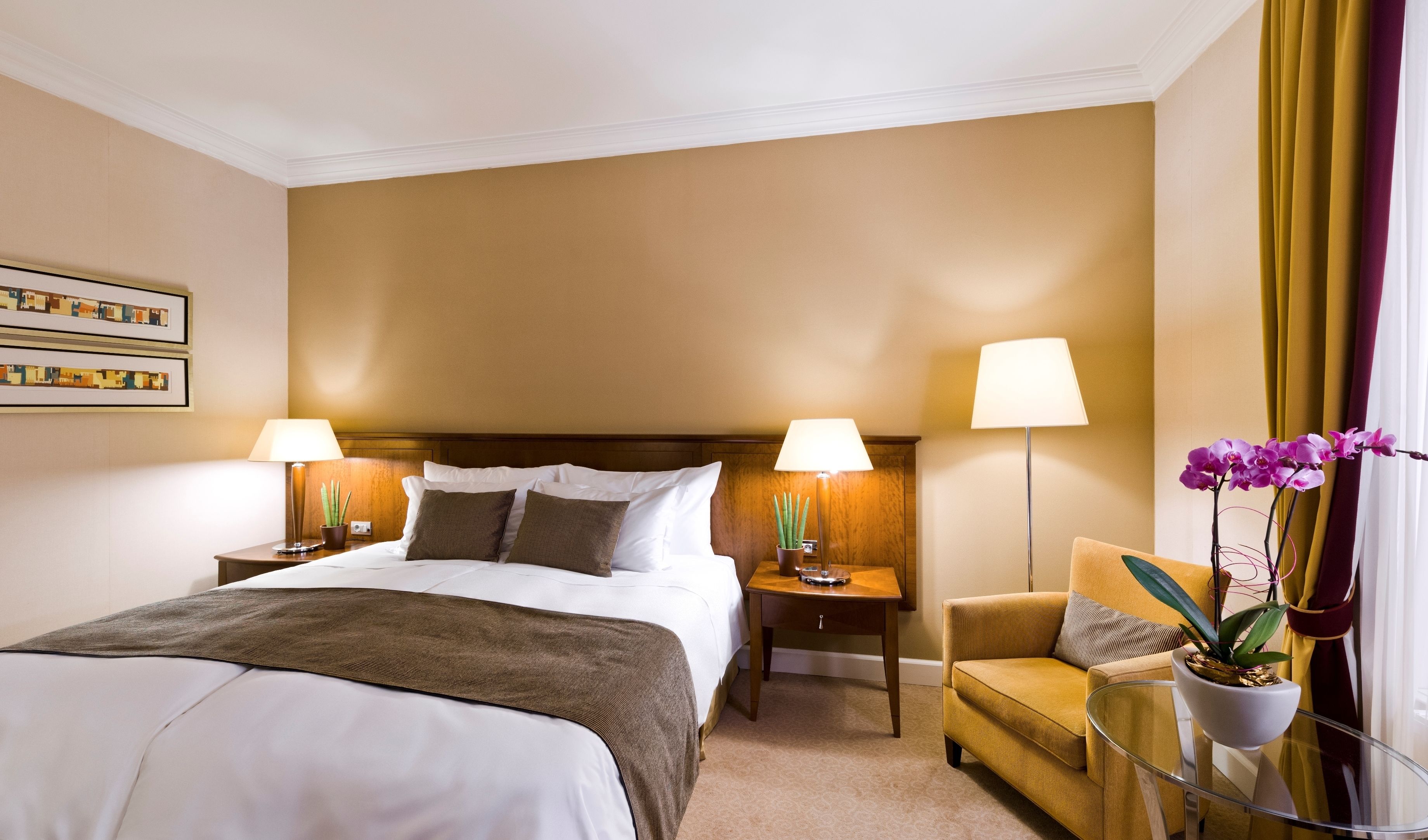 Una habitación de hotel con una cama grande, dos mesitas de noche con lámparas, un pequeño sofá y una orquídea en maceta.