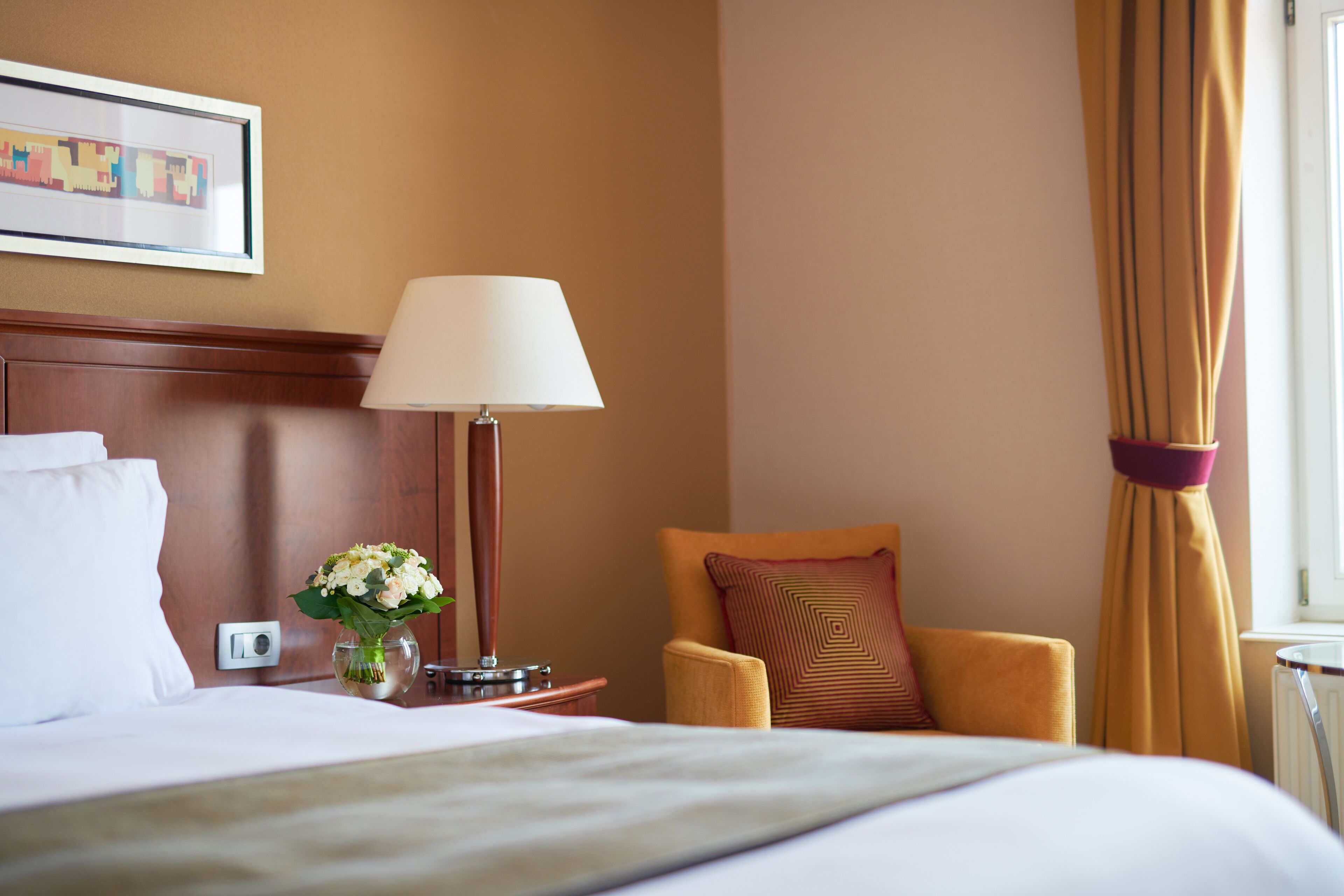 Habitación de hotel con cama, mesita de noche con lámpara, un jarrón con flores, una silla con cojín y ventana con cortinas.