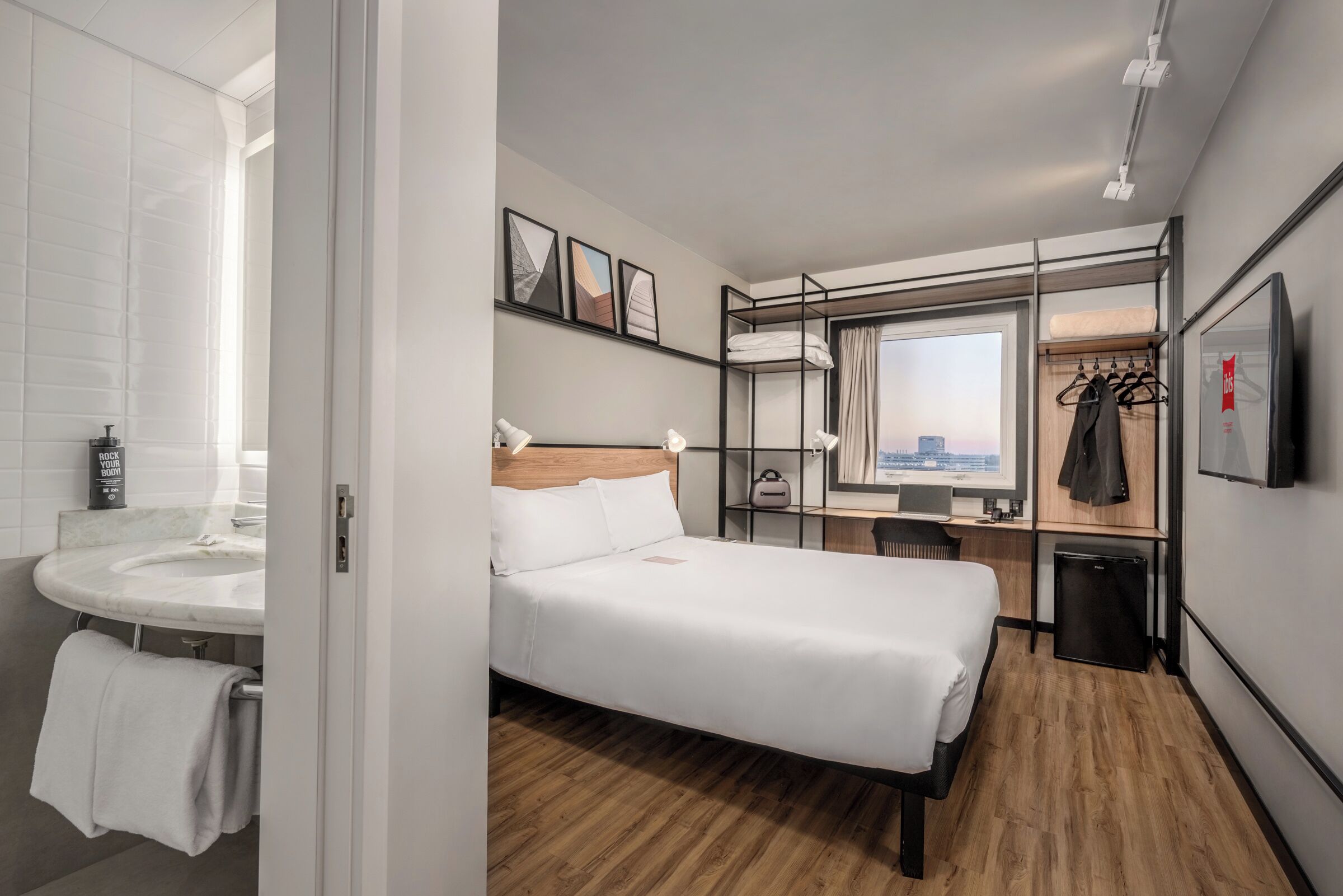 Une chambre d’hôtel équipée d’un lit, d’un bureau, d’une chaise, d’un petit réfrigérateur, d’un miroir et d’une fenêtre donnant sur la ville.