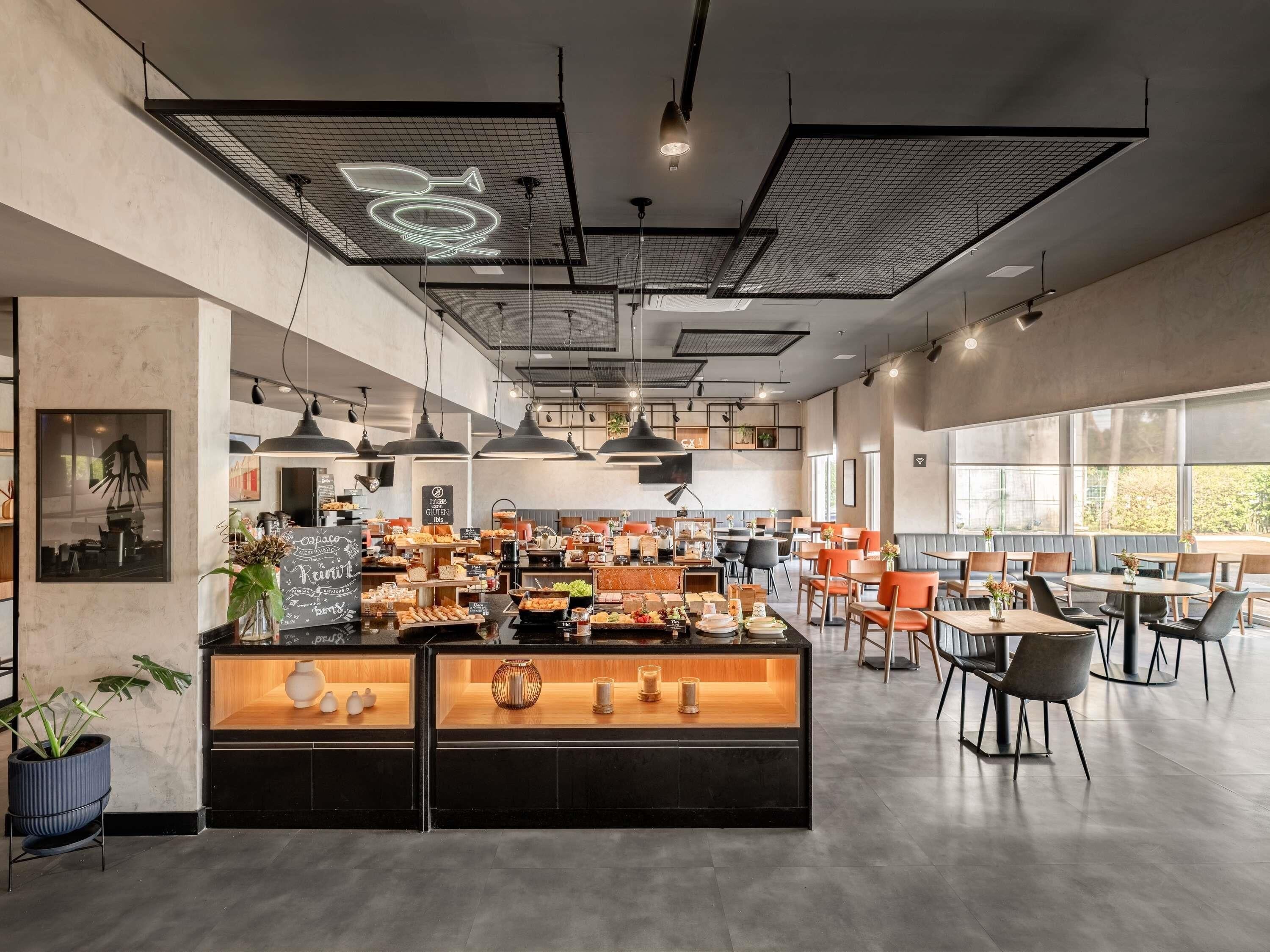 Un café moderno con una barra central que exhibe diferentes productos alimenticios, varias zonas de estar con mesas y sillas, y un gran ventanal que proporciona luz natural.