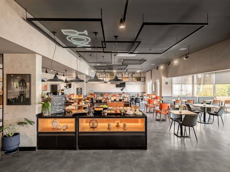 A modern cafe interior with a central counter displaying various food items, multiple seating areas with tables and chairs, and a large window providing natural light.