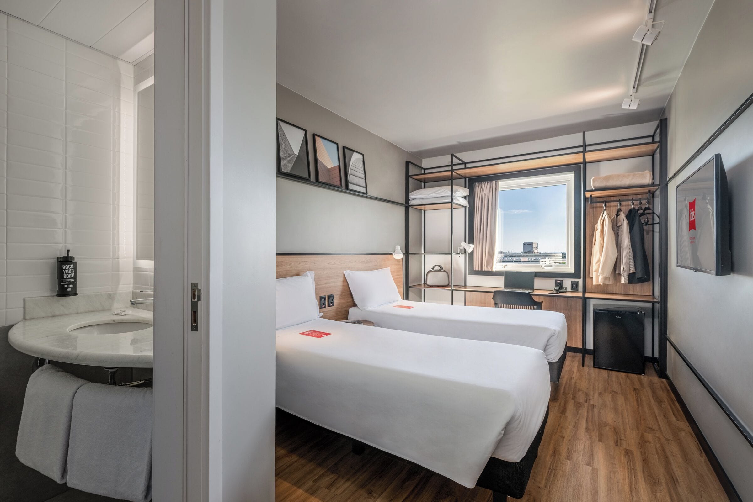 A hotel room with two beds, a desk, a small fridge, and a view of the city.