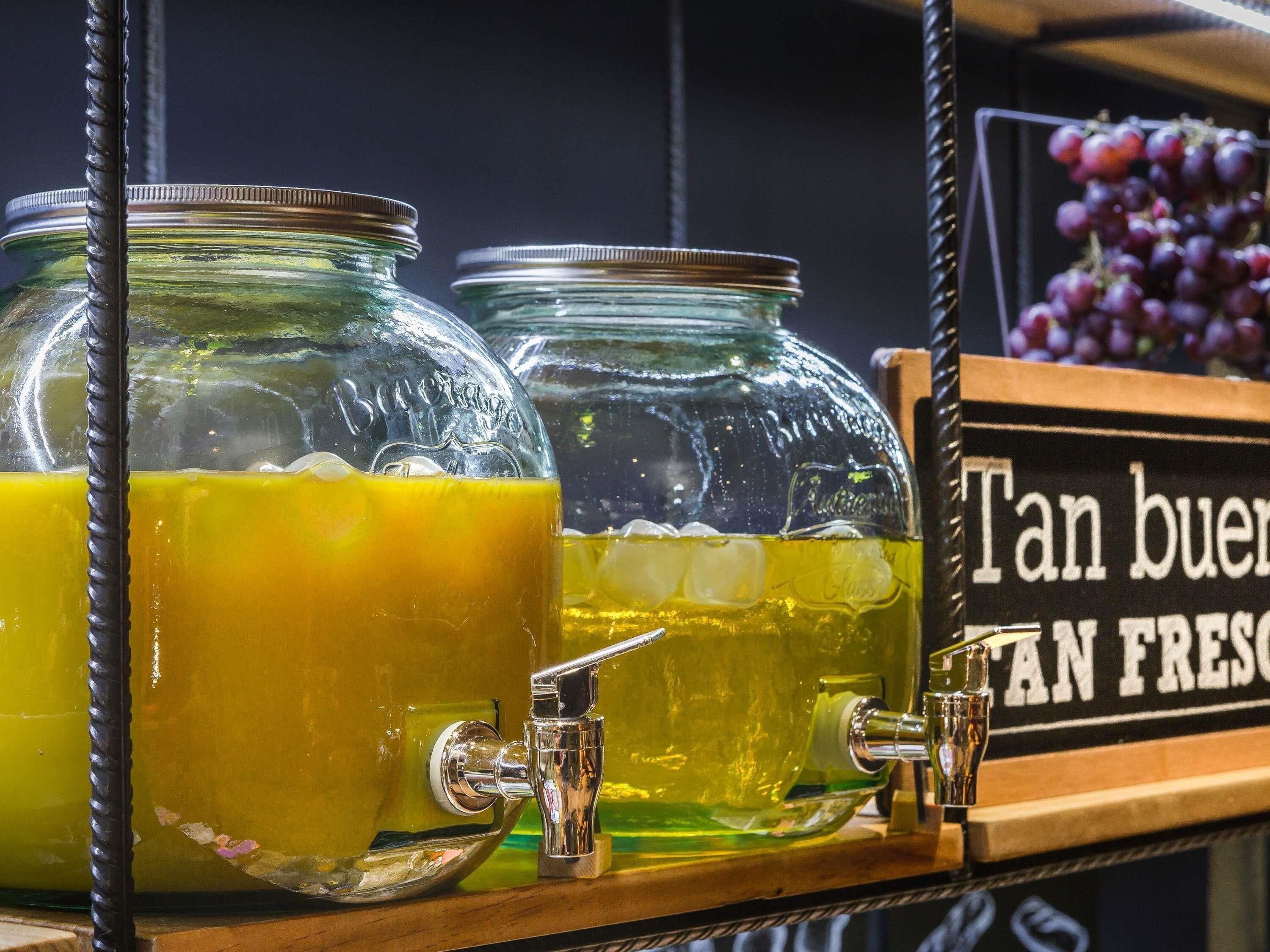 Un dispensador de bebidas con tres frascos de vidrio que contienen líquidos, un grifo metálico y un cartel con texto.