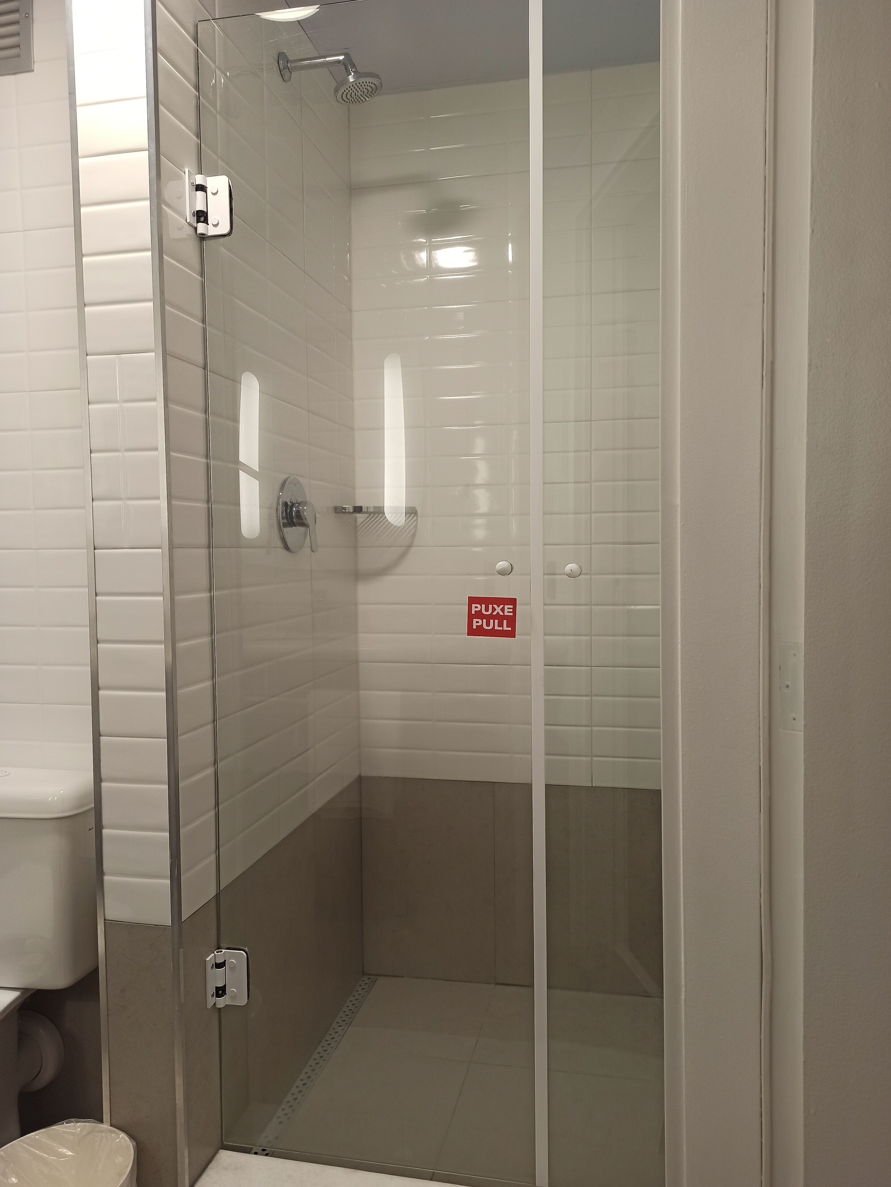 Un box de ducha con paredes de azulejos blancos, un secamanos visible y un cartel que dice 'PUXE PULL'.
