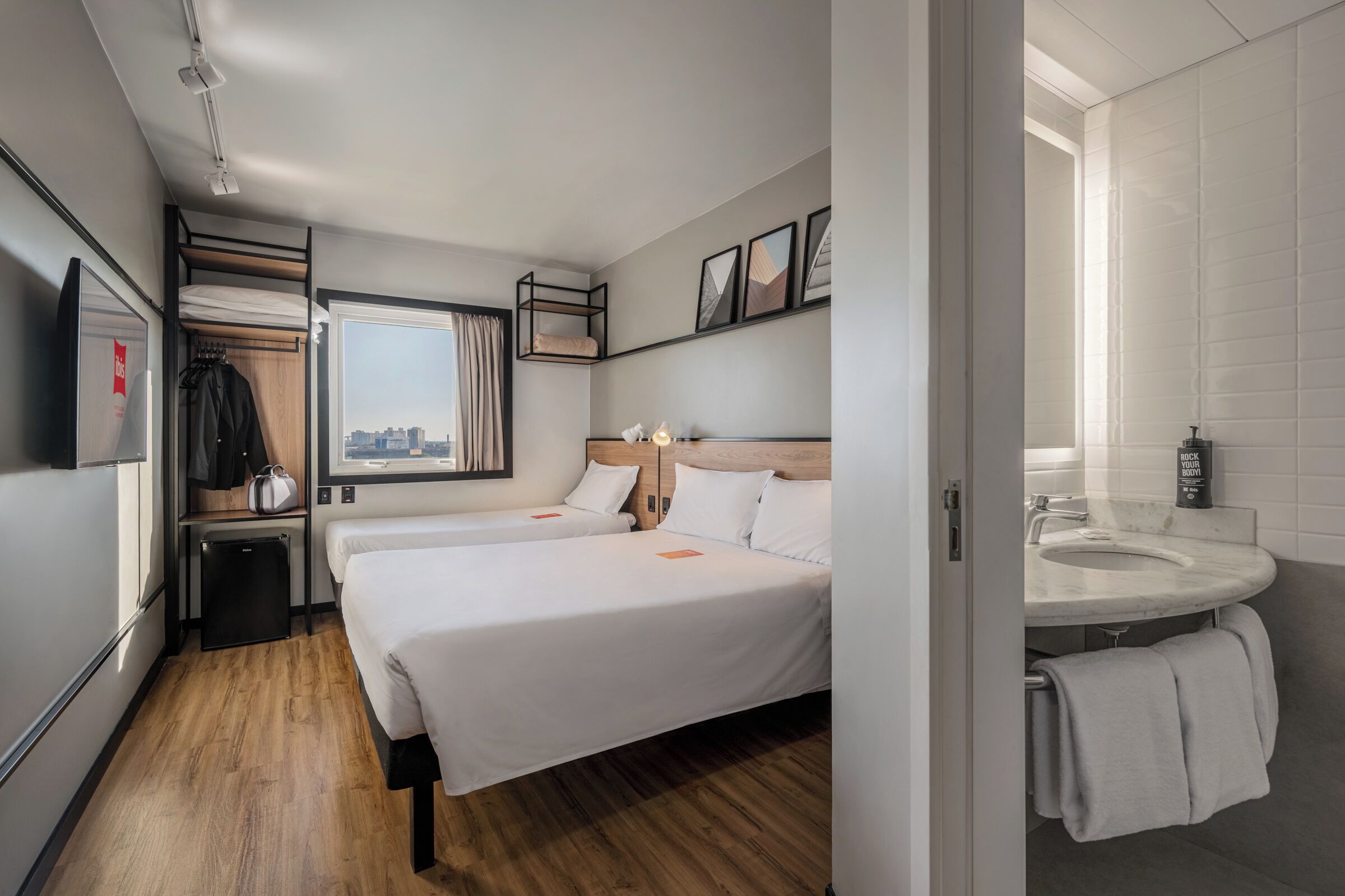 Habitación de hotel con cama, mesita de noche, ventana con vista a la ciudad, armario, refrigerador pequeño y baño con lavamanos y toalla.