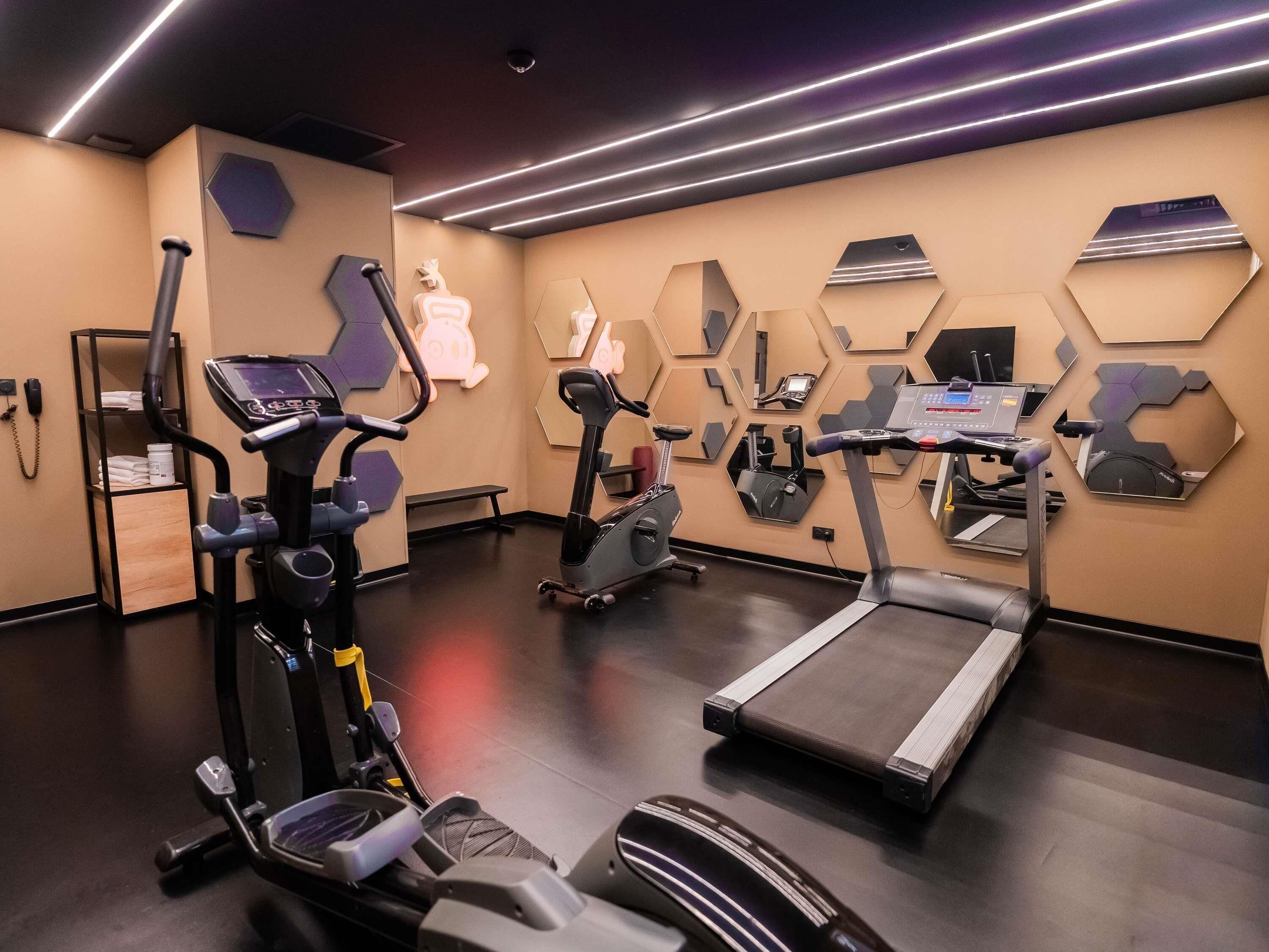 Un gimnasio con varios equipos de ejercicio, incluyendo cintas de correr y bicicletas estáticas, y una pared con espejos hexagonales.